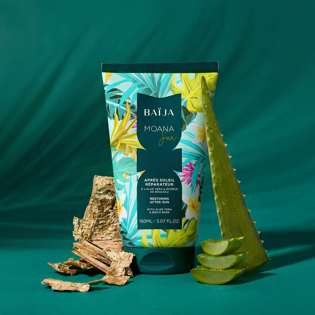 Tube “BAÏJA MOANA Sun – Après Soleil Réparateur” placé debout au centre, sur un fond vert/vert-turquoise. À droite, une grande feuille d’aloe vera dressée, avec des gouttes d’eau, et plusieurs tranches d’aloe au premier plan. À gauche, des morceaux d’écorce/bois (aspect écorce de bouleau) disposés en tas. Éclairage studio avec ombres nettes, mise en scène “ingrédients naturels”.