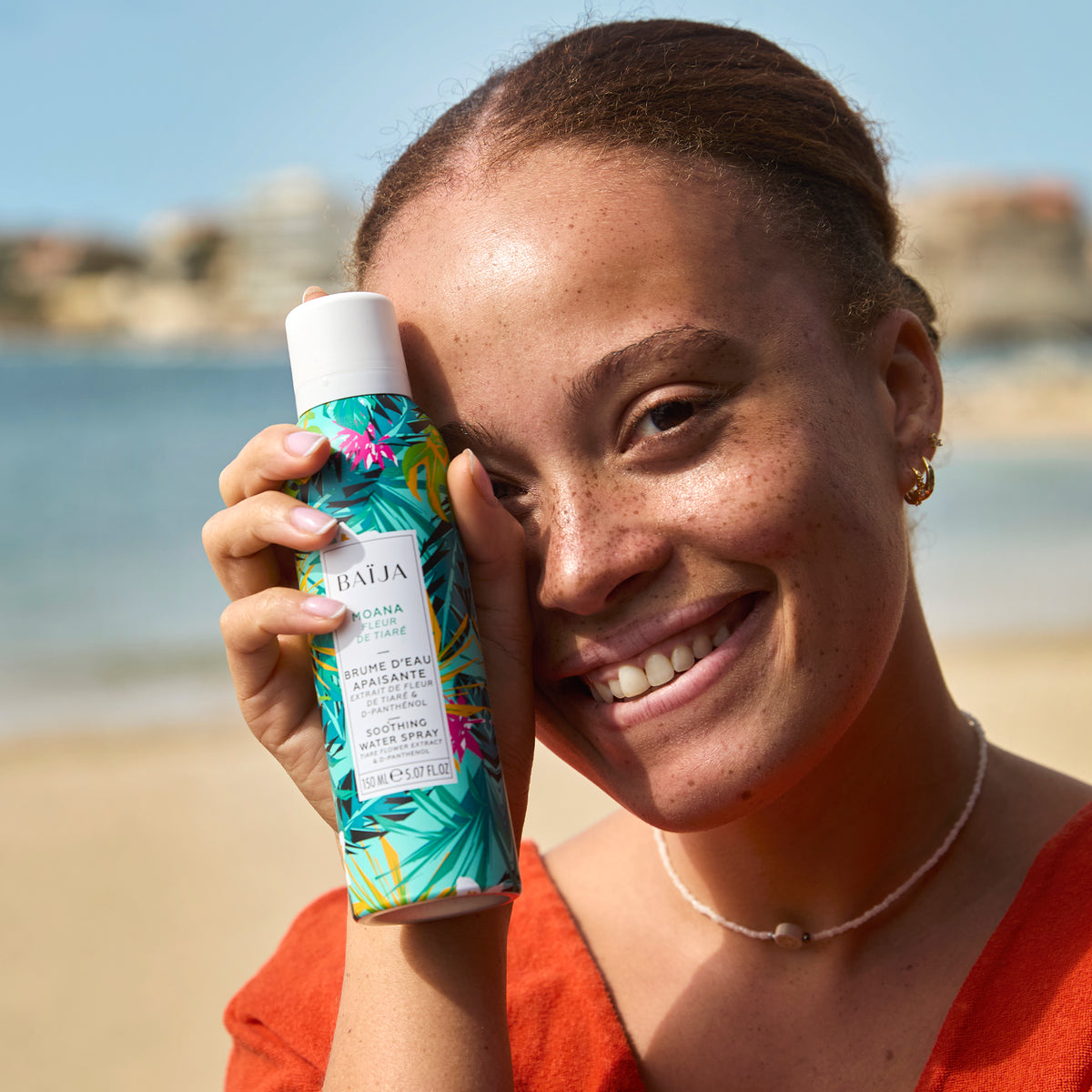 Femme souriante tenant la brume d’eau apaisante Moana près de son visage sur la plage, incarnant la fraîcheur et la joie de la collection solaire BAÏJA.