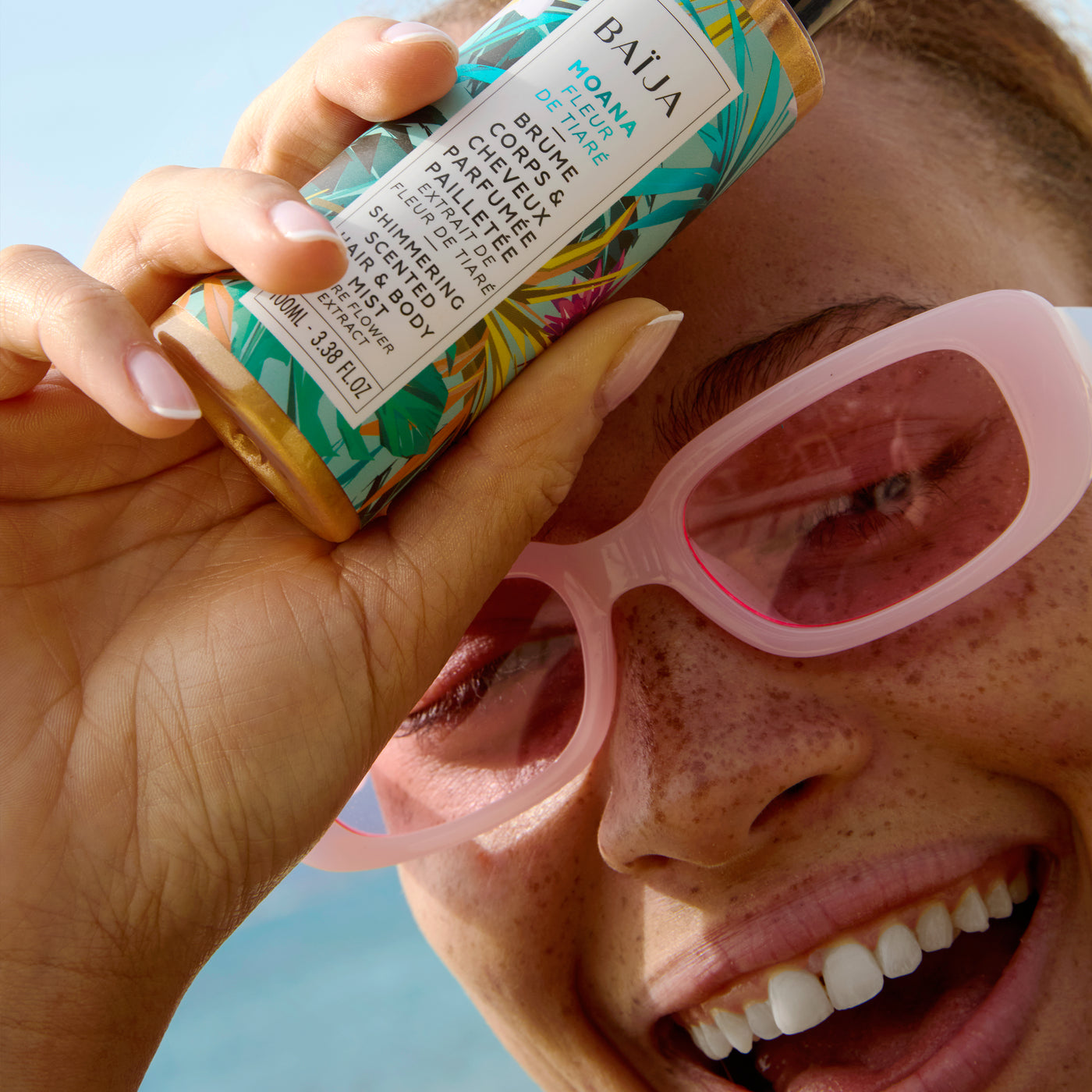 Femme souriante portant des lunettes roses et tenant la brume corps & cheveux parfumée pailletée Moana, dans une ambiance solaire et joyeuse.