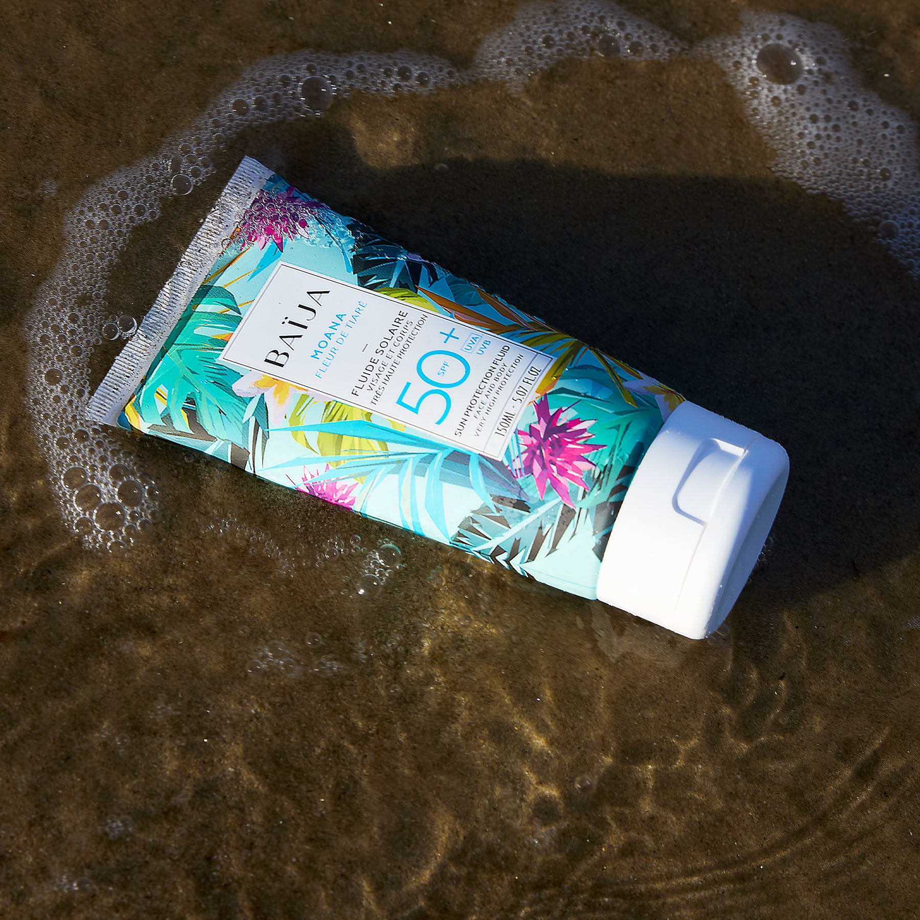 Tube du fluide solaire Moana SPF 50+  posé sur le sable au bord de l’eau, packaging tropical coloré légèrement entouré d’écume.