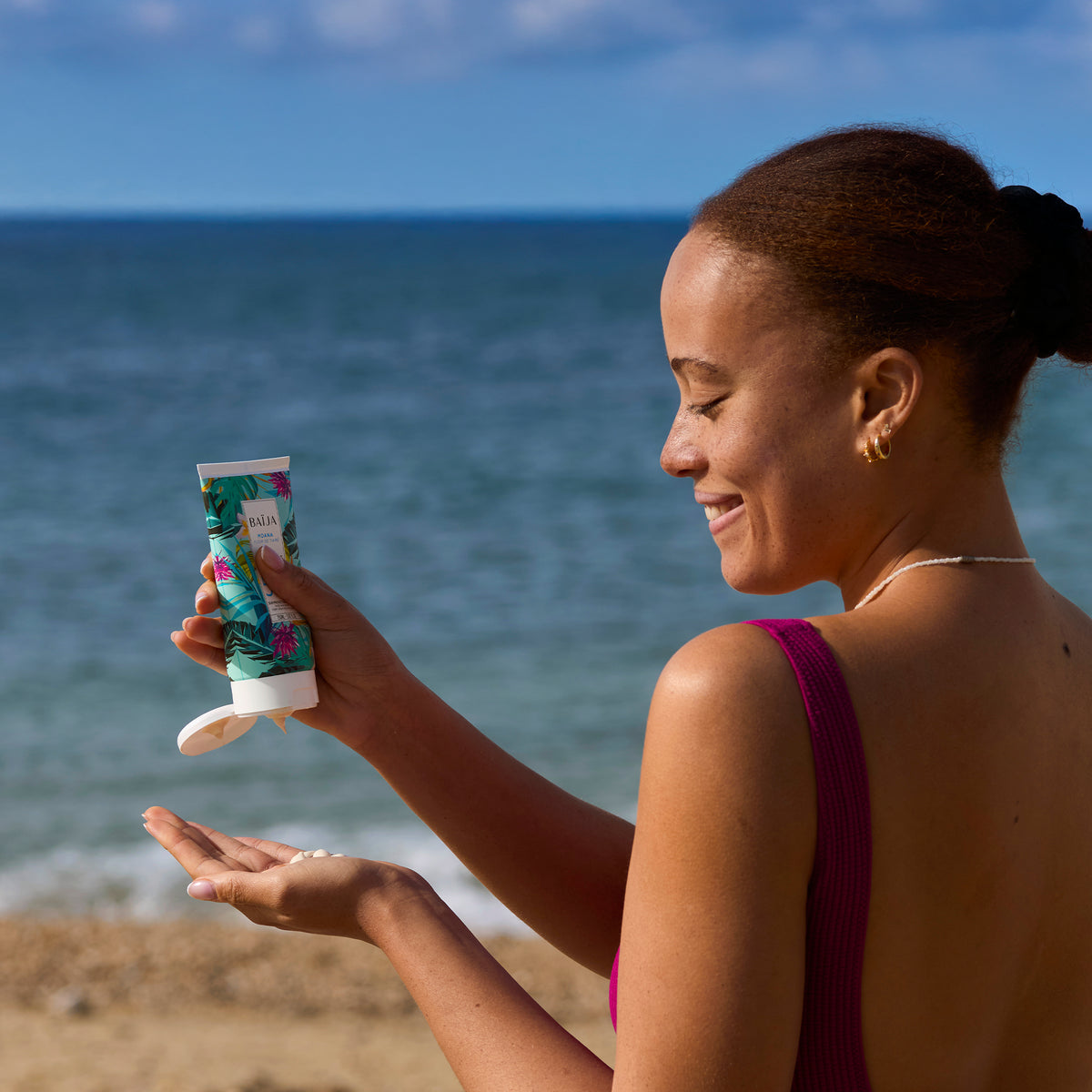 Femme souriante appliquant le fluide solaire SPF 50+Moana sur la main, en bord de mer, vêtue d’un maillot prune.