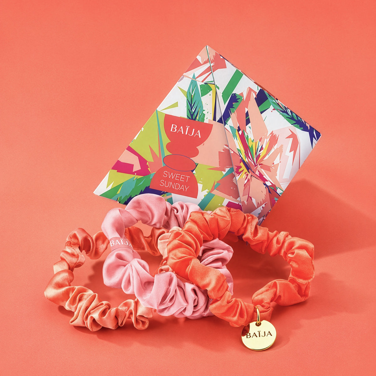 Ensemble de chouchous satinés Baïja Sweet Sunday aux teintes rose poudré et corail, présenté avec sa pochette graphique colorée