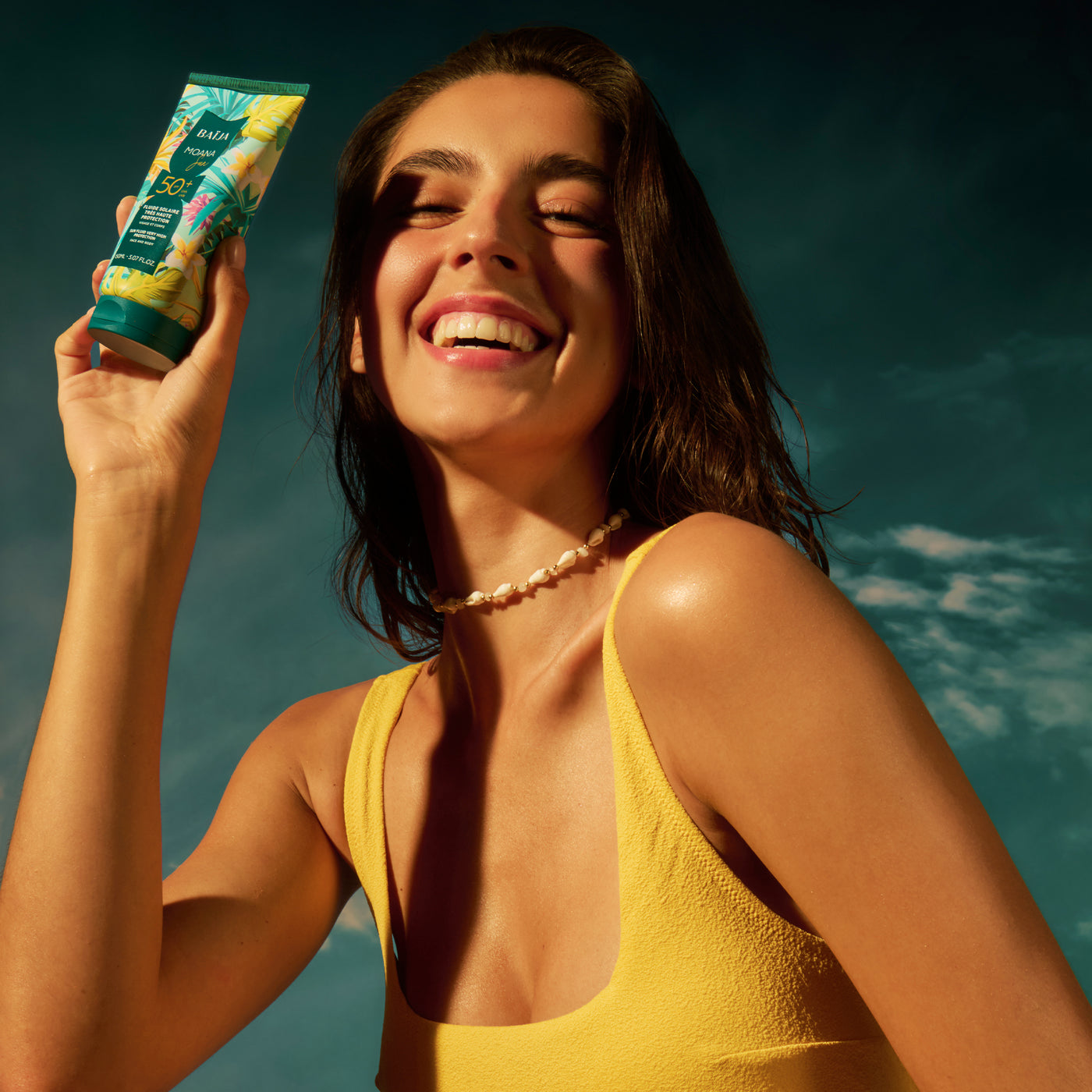 Jeune femme rayonnante sous une lumière solaire tenant un fluide solaire SPF50+ Baïja Moana Sun, peau glowy protégée UVA UVB