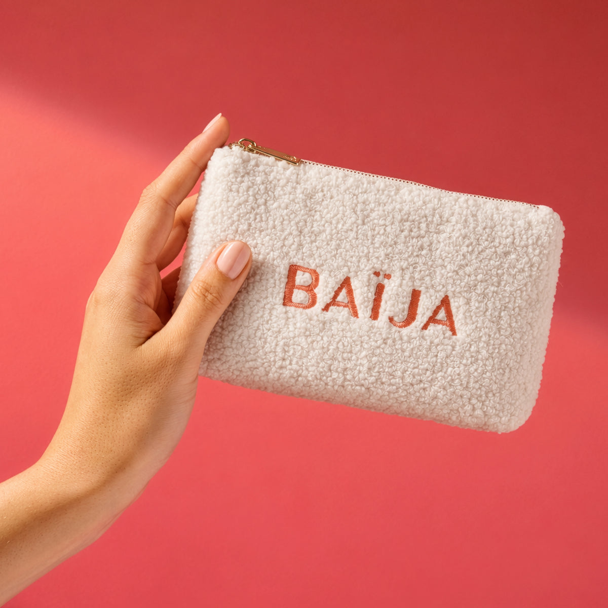 Trousse zippée Baïja en bouclette écrue avec logo brodé, présentée à la main dans une ambiance lumineuse rose