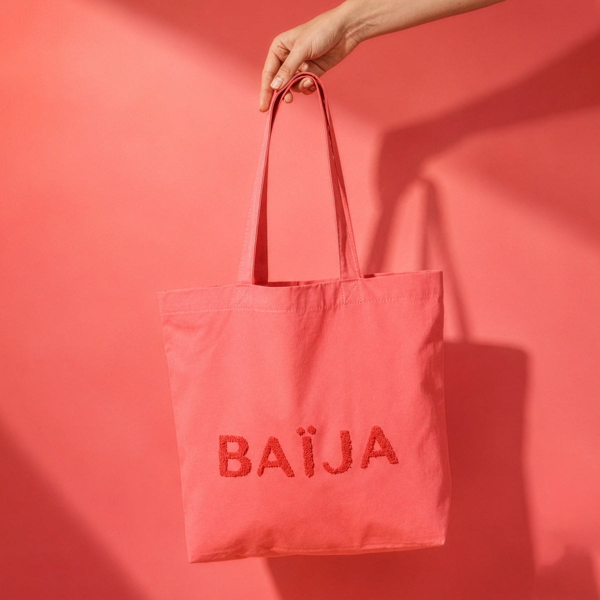 Grand tote bag rose Baïja en coton avec logo texturé, suspendu à la main dans une ambiance solaire aux tons corail