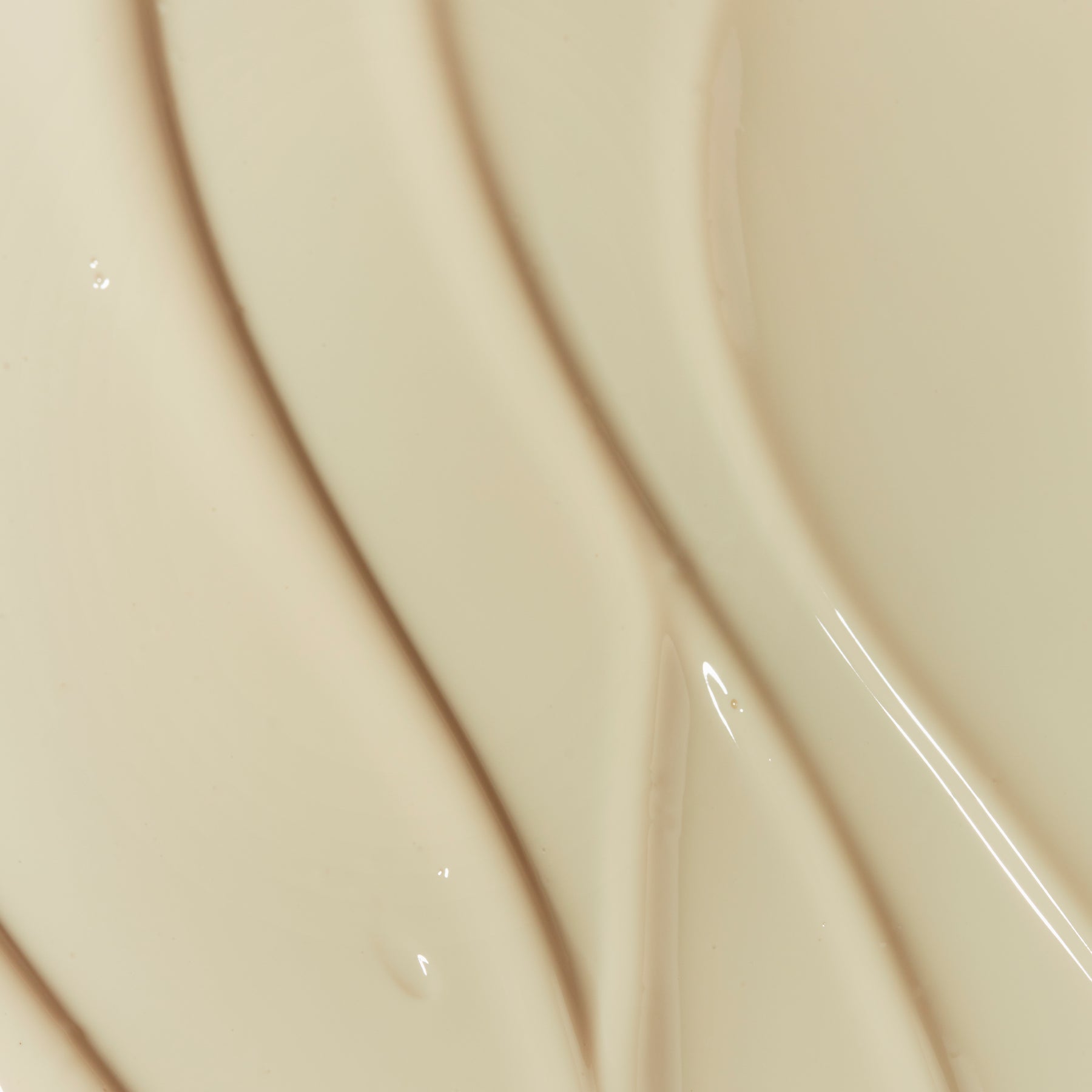 Gros plan abstrait d’une texture crème/beige claire, lisse et brillante. On voit des traînées courbes comme des coups de spatule et quelques reflets lumineux sur la surface. Aucun produit ni texte visible, uniquement la matière.