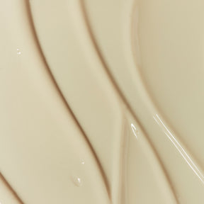 Gros plan abstrait d’une texture crème/beige claire, lisse et brillante. On voit des traînées courbes comme des coups de spatule et quelques reflets lumineux sur la surface. Aucun produit ni texte visible, uniquement la matière.