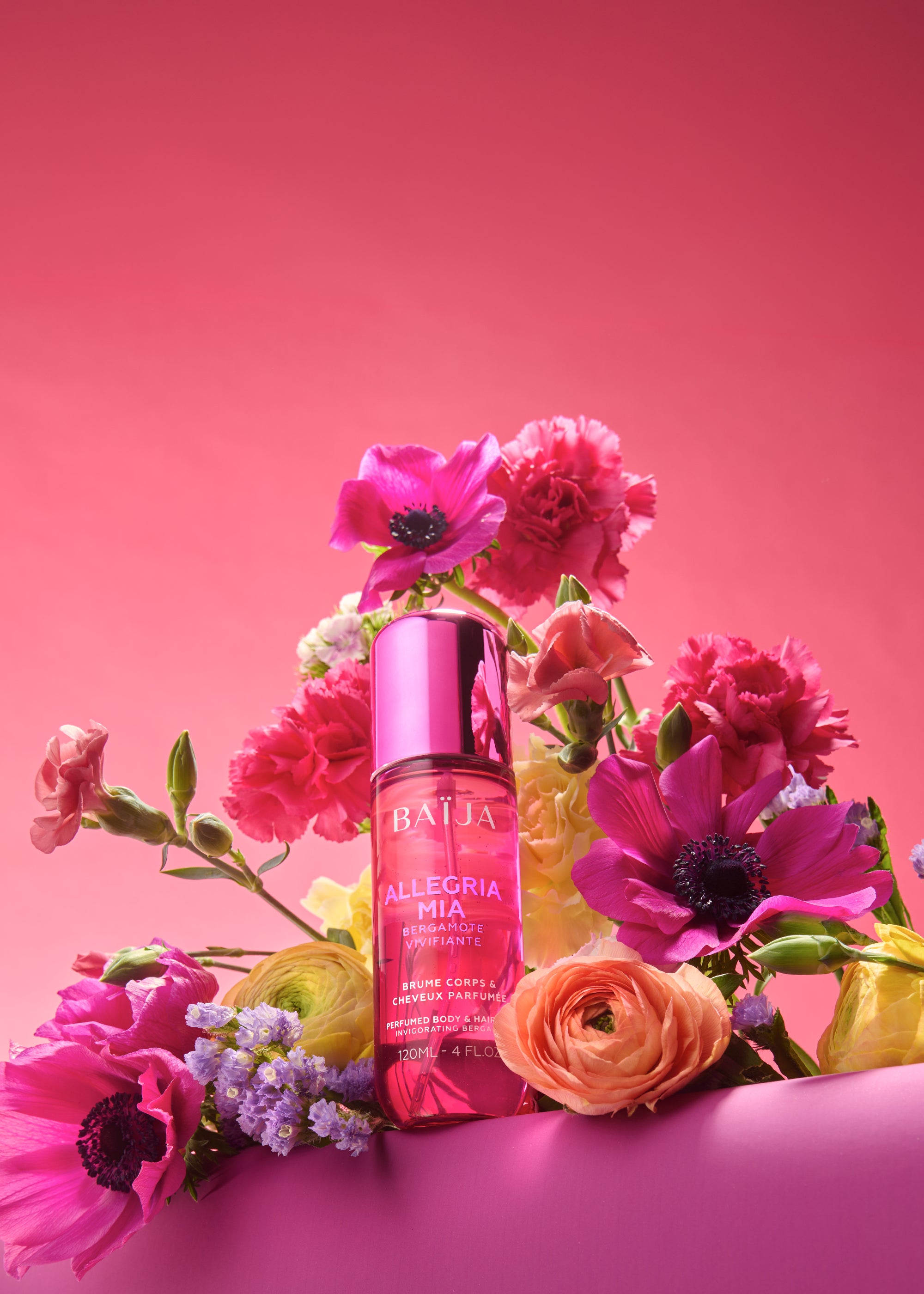 Flacon rose Baïja Allegria Mia (brume corps et cheveux) posé devant un bouquet de fleurs fuchsia, corail et jaunes, sur fond rose.