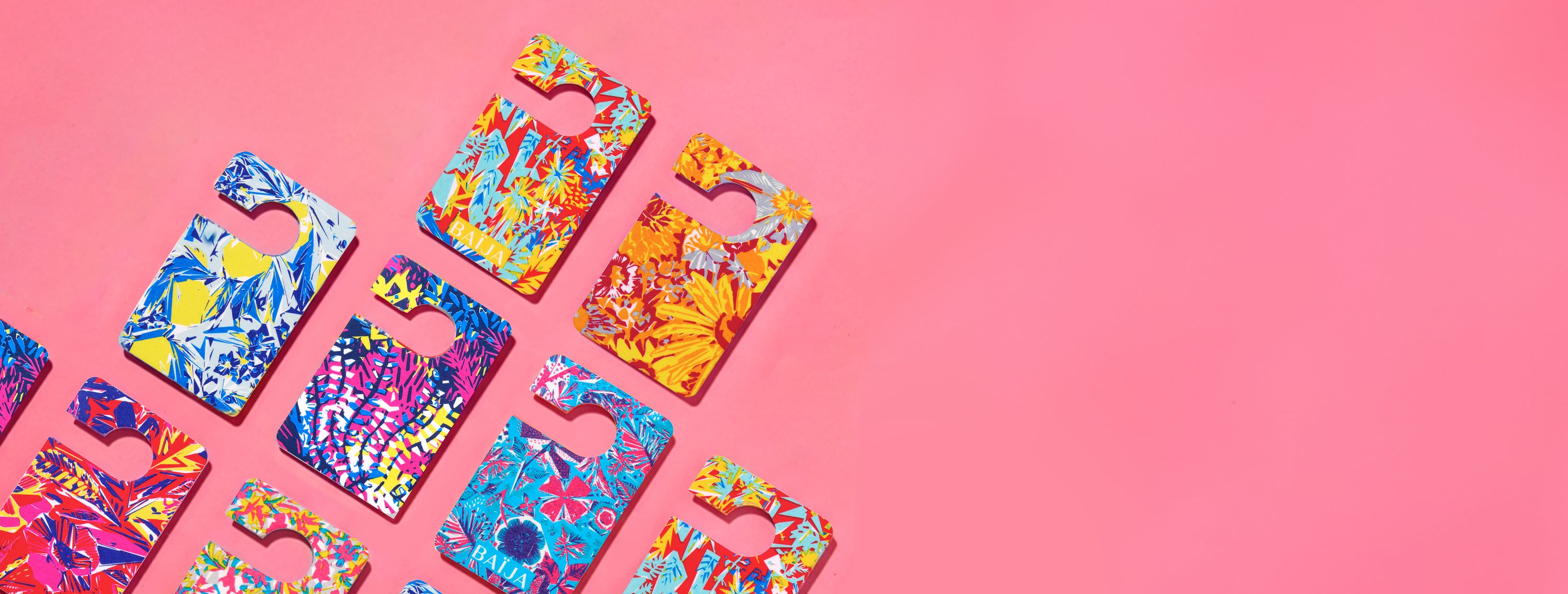 Flatlay de cartes de parfums voiture Baïja aux imprimés tropicaux et graphiques, disposées en diagonale sur un fond rose uni.