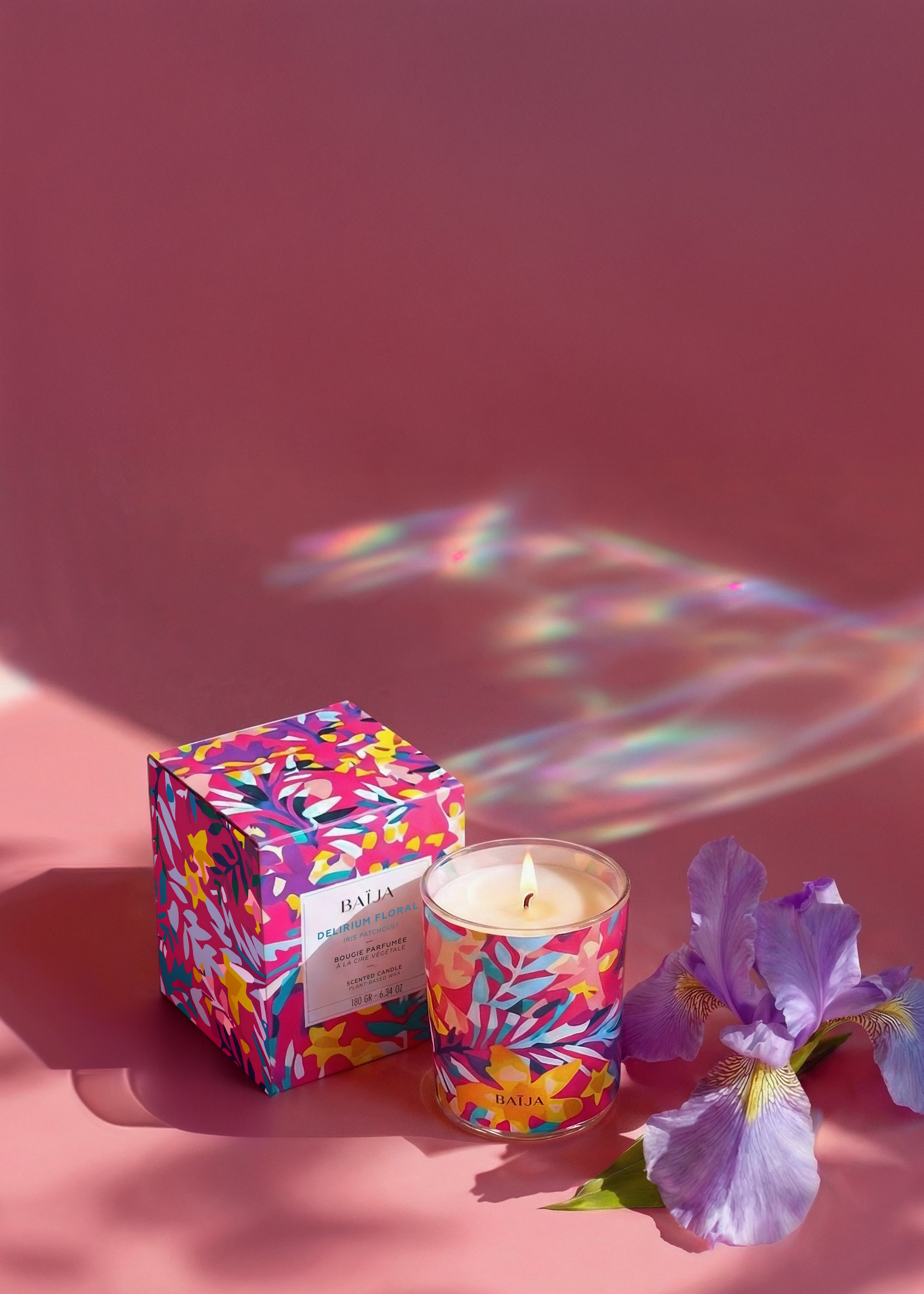 Bougie parfumée Baïja Delirium Floral dans un verre illustré multicolore, avec sa boîte assortie, posée sur fond rose à côté d’un iris violet, reflets lumineux irisés.