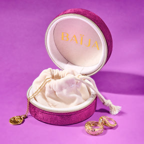 Boîte à bijoux ronde en velours fuchsia ouverte sur fond violet, contenant une pochette crème et des bijoux dorés à l'intérieur. 