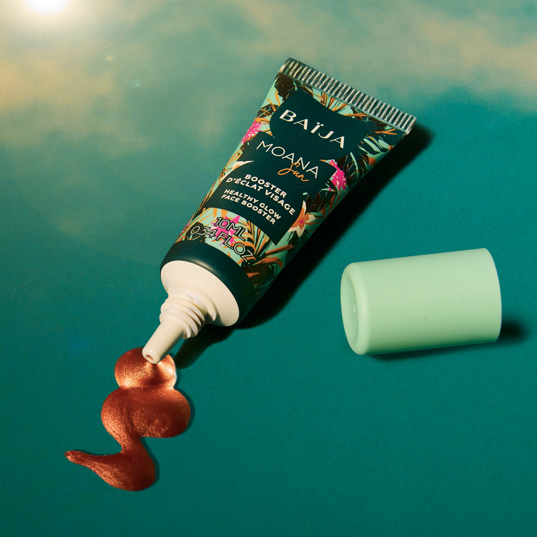 Gros plan en studio sur fond dégradé vert/bleu avec effet de soleil et lens flare en haut à gauche. Le tube “BAÏJA Moana Sun” est couché en diagonale, capuchon vert menthe séparé à droite. À l’embout, une noisette de matière liquide/cuivrée est déposée sur la surface.