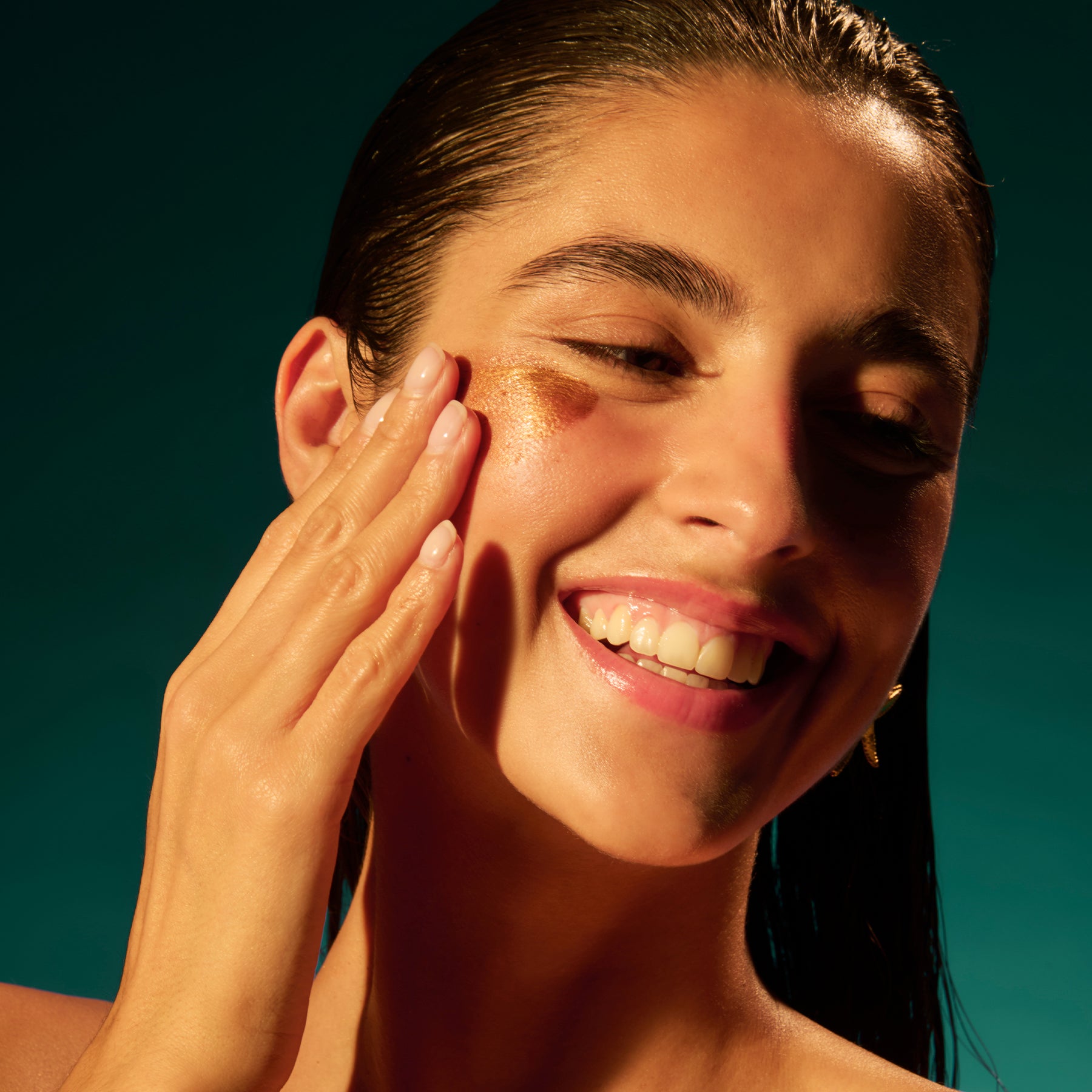 Gros plan sur une joue illuminée par une touche de texture nacrée dorée, effet “glow” très réfléchissant. La matière est étalée en un trait dense et crémeux, avec un fini métallique lumineux qui capte fortement la lumière. Une main aux ongles nude applique la texture sur la pommette ; le modèle sourit, sur fond uni bleu-vert, avec une lumière chaude et contrastée.