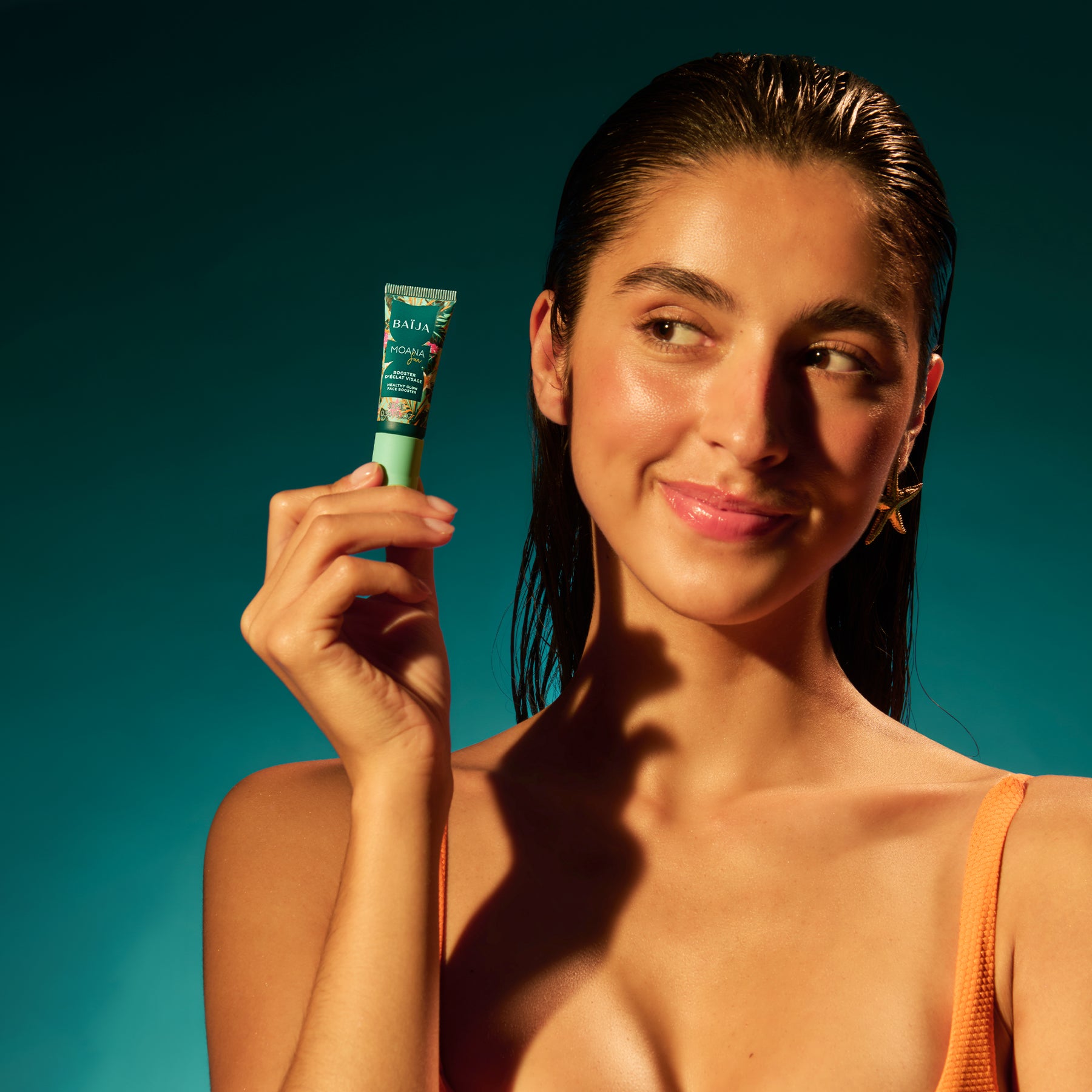 Portrait en studio sur fond vert/bleu : une femme (cheveux mouillés) tient le petit tube BAÏJA près de sa joue, en regardant légèrement sur le côté. Éclairage très contrasté type soleil, ombres marquées sur le visage et le cou. Maillot orange visible.