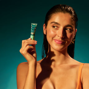 Portrait en studio sur fond vert/bleu : une femme (cheveux mouillés) tient le petit tube BAÏJA près de sa joue, en regardant légèrement sur le côté. Éclairage très contrasté type soleil, ombres marquées sur le visage et le cou. Maillot orange visible.