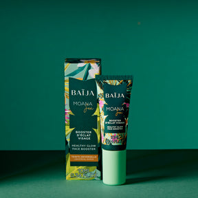Photo produit en studio sur fond vert/bleu pétrole uni. Un petit tube cosmétique “BAÏJA Moana Sun” est posé debout, avec son étui carton juste derrière. Le tube a un habillage tropical multicolore (feuillages/fleurs) et un capuchon vert menthe. Éclairage dur avec ombres marquées au sol.