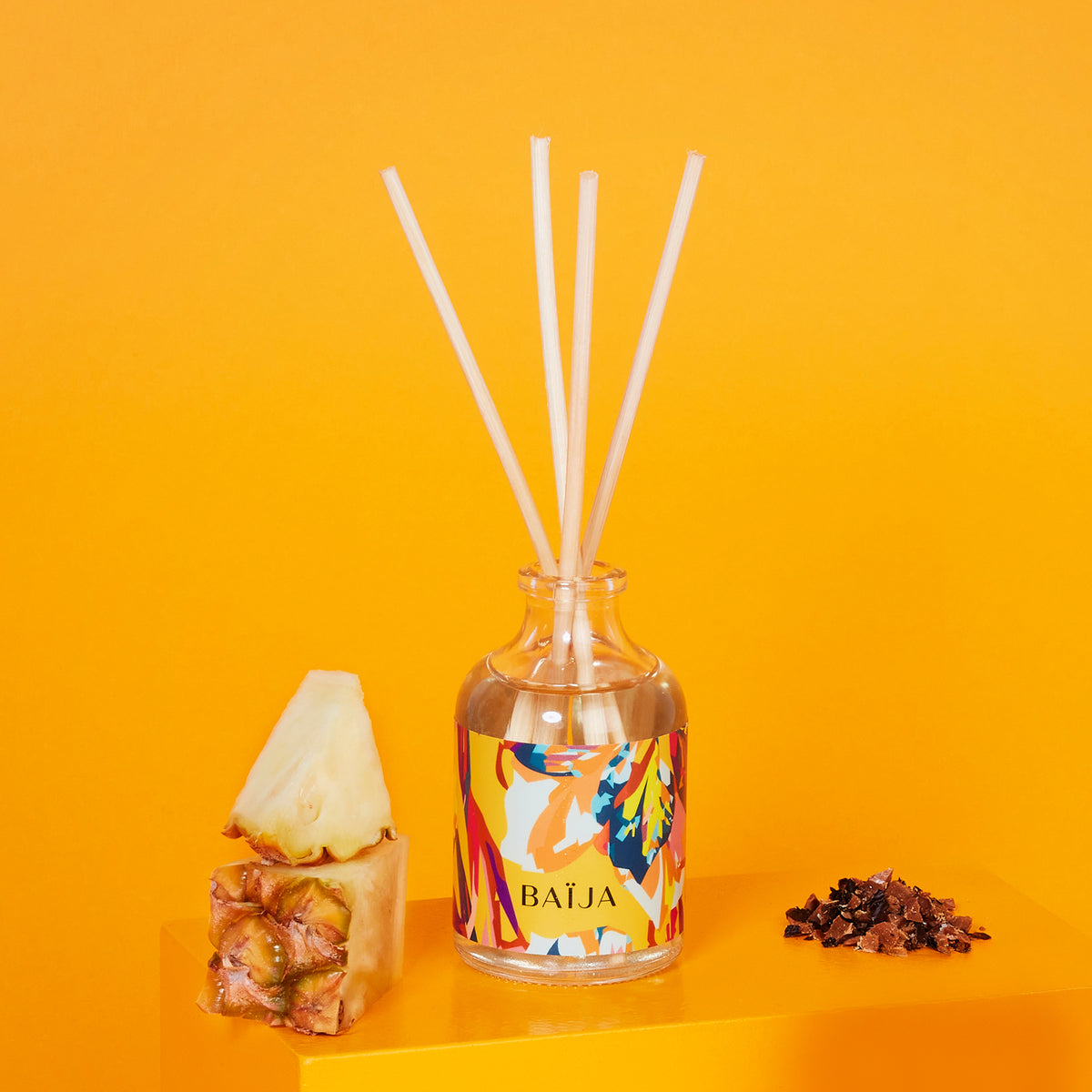Bouquet parfumé BAÏJA Lost Paradise avec bâtonnets diffuseurs, accompagné d’un morceau d’ananas frais et d’éclats de fève tonka sur un fond jaune solaire.
