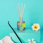 Bouquet parfumé BAÏJA Moana avec bâtonnets diffuseurs, entouré d’une gousse de vanille, d’un morceau de noix de coco et de fleurs de tiaré sur un fond turquoise lumineux.
