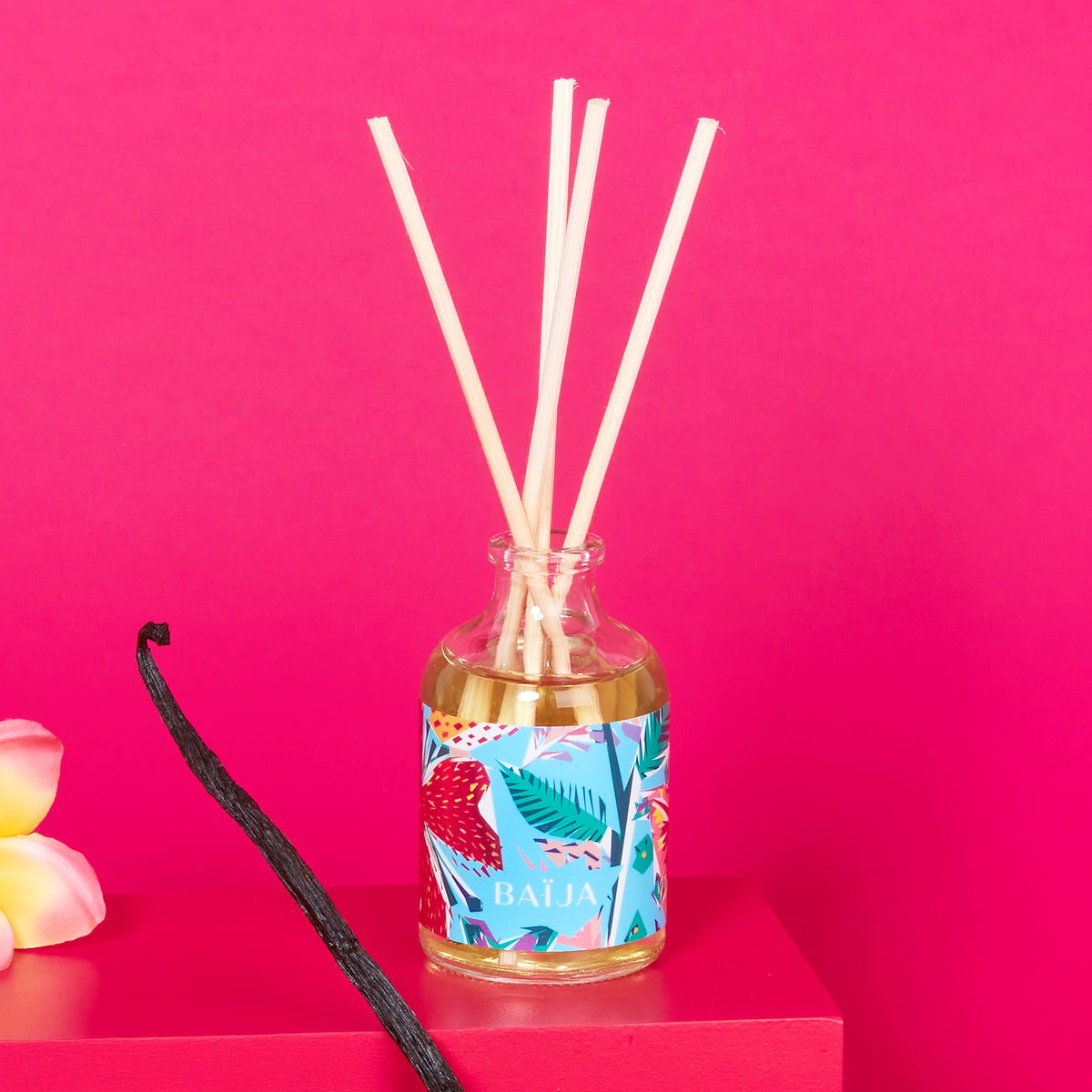 Bouquet parfumé BAÏJA Île d’Azur avec bâtonnets diffuseurs, accompagné d’une gousse de vanille et d’une fleur de tiaré sur fond rose vif.