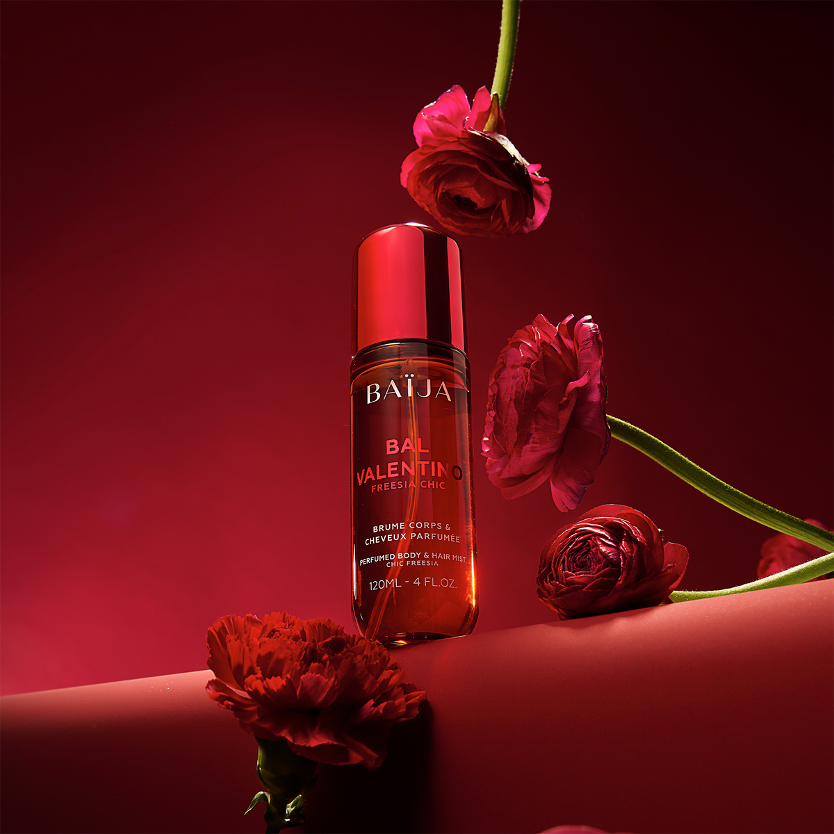 Flacon Baïja Bal Valentino brume parfumée, posé sur un décor rouge avec fleurs rouges en suspension et au premier plan, lumière chaude.