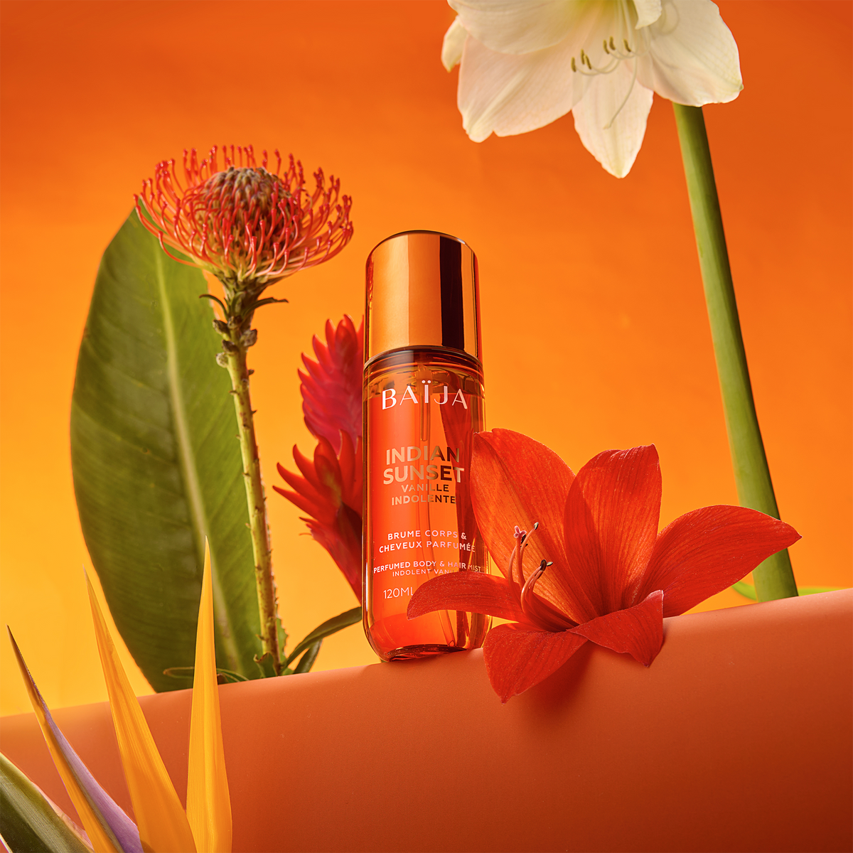 Flacon Baïja Indian Sunset brume parfumée corps et cheveux, posé parmi fleurs oranges et blanches sur décor orange éclatant.