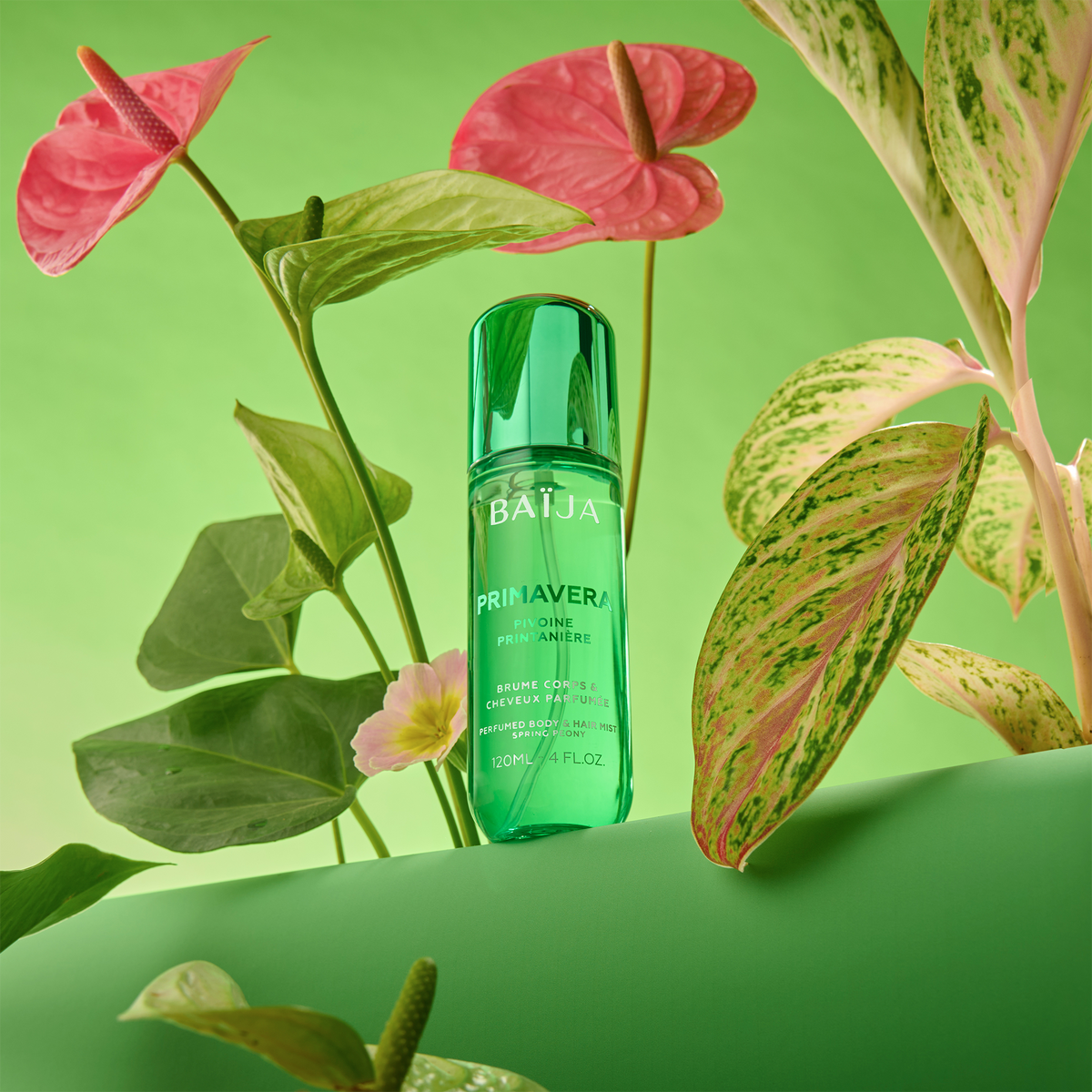 Flacon Baïja Primavera brume parfumée corps et cheveux, posé parmi feuilles tropicales et fleurs roses, ambiance verte lumineuse.