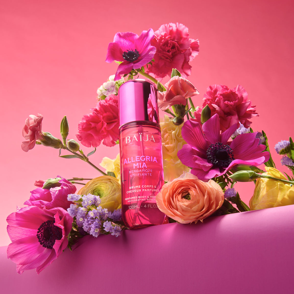 Flacon rose Baïja Allegria Mia brume parfumée, mis en scène avec fleurs fuchsia, corail et jaunes sur décor rose.
