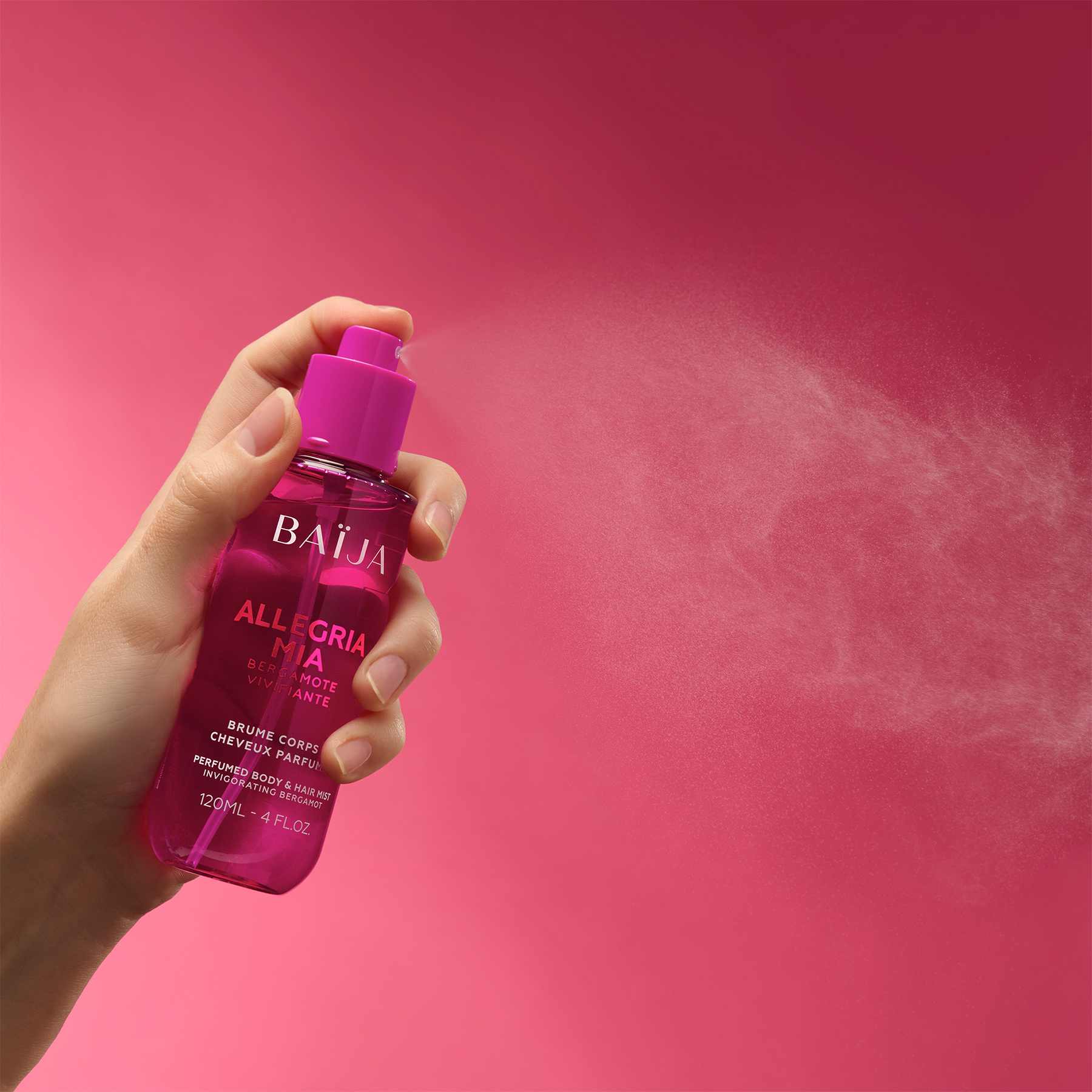 Main tenant le spray Baïja Allegria Mia et diffusant une fine brume dans l’air, décor rose dégradé.