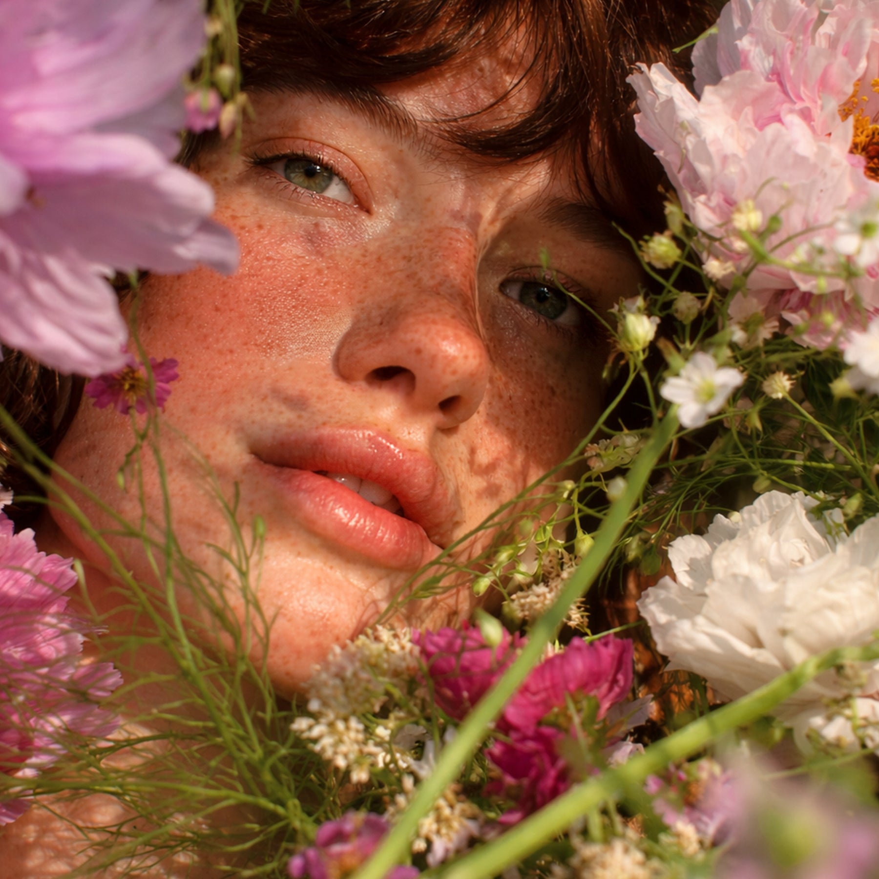Visage cadré serré parmi des fleurs roses et blanches, lumière douce, regard direct vers l’objectif.