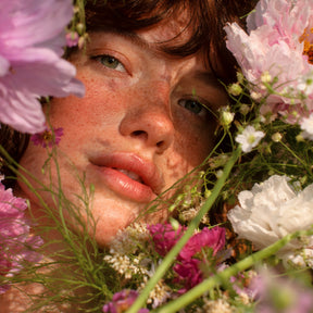 Visage cadré serré parmi des fleurs roses et blanches, lumière douce, regard direct vers l’objectif.