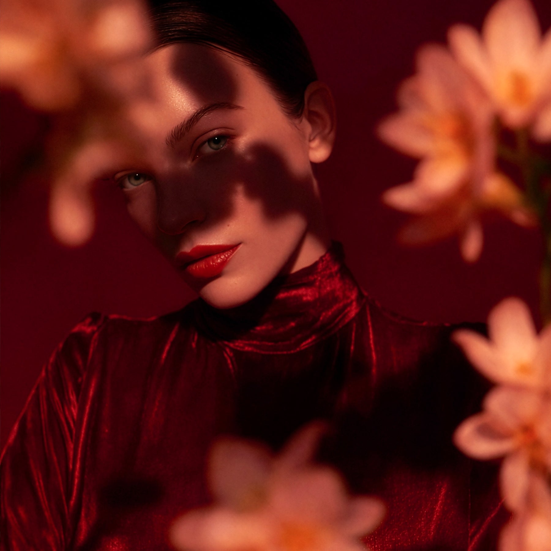 Gros plan d’une femme en haut col rouge, lumière tamisée et silhouettes de fleurs floutées au premier plan.