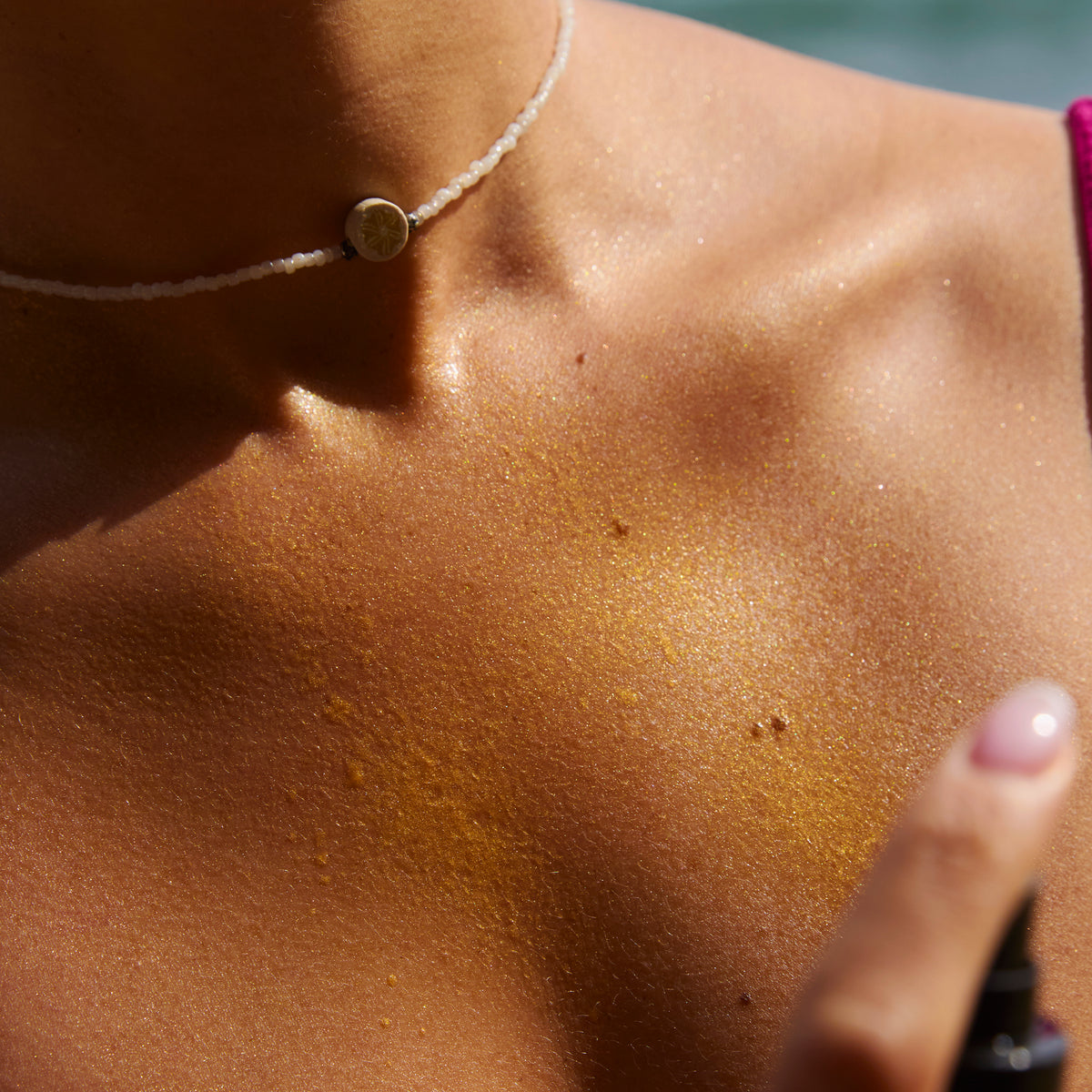 Photo lifestyle : gros plan d’un buste/épaule/haut du torse d’une personne en extérieur ou sous lumière solaire, peau brillante avec des micro-paillettes dorées visibles sur la peau. Arrière-plan flou (tons bleus/verts). Le produit n’est pas visible, l’image met l’accent sur l’effet “glow” et la texture pailletée.