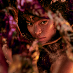 Visage au centre d’une composition florale sombre, lumière chaude, fumée légère et fleurs floutées autour.