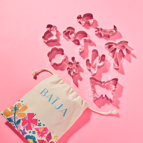 Emporte-pièces métalliques de formes variées sortant d’un sachet en coton décoré de motifs floraux colorés, sur fond rose vif, provenant de la collection Île d’Azur. 