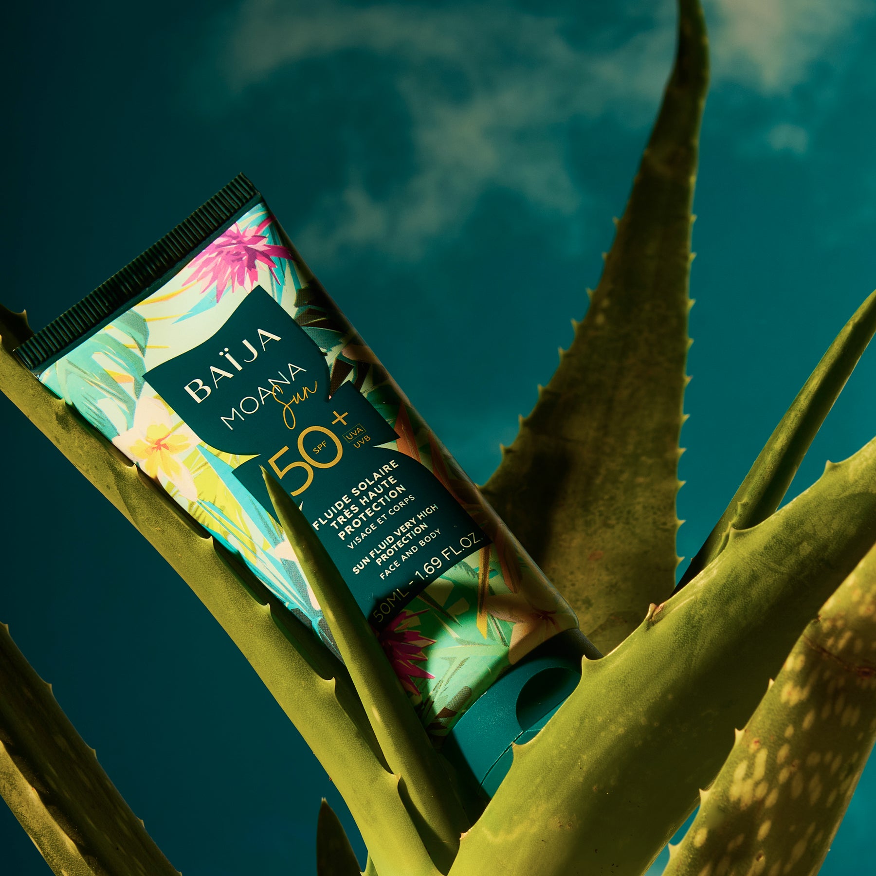 Tube de crème solaire Baïja Moana Sun SPF 50+ en gros plan, posé en biais au milieu de grandes feuilles d’aloe vera. Fond bleu-vert avec lumière dramatique et ombres marquées, ambiance tropicale.