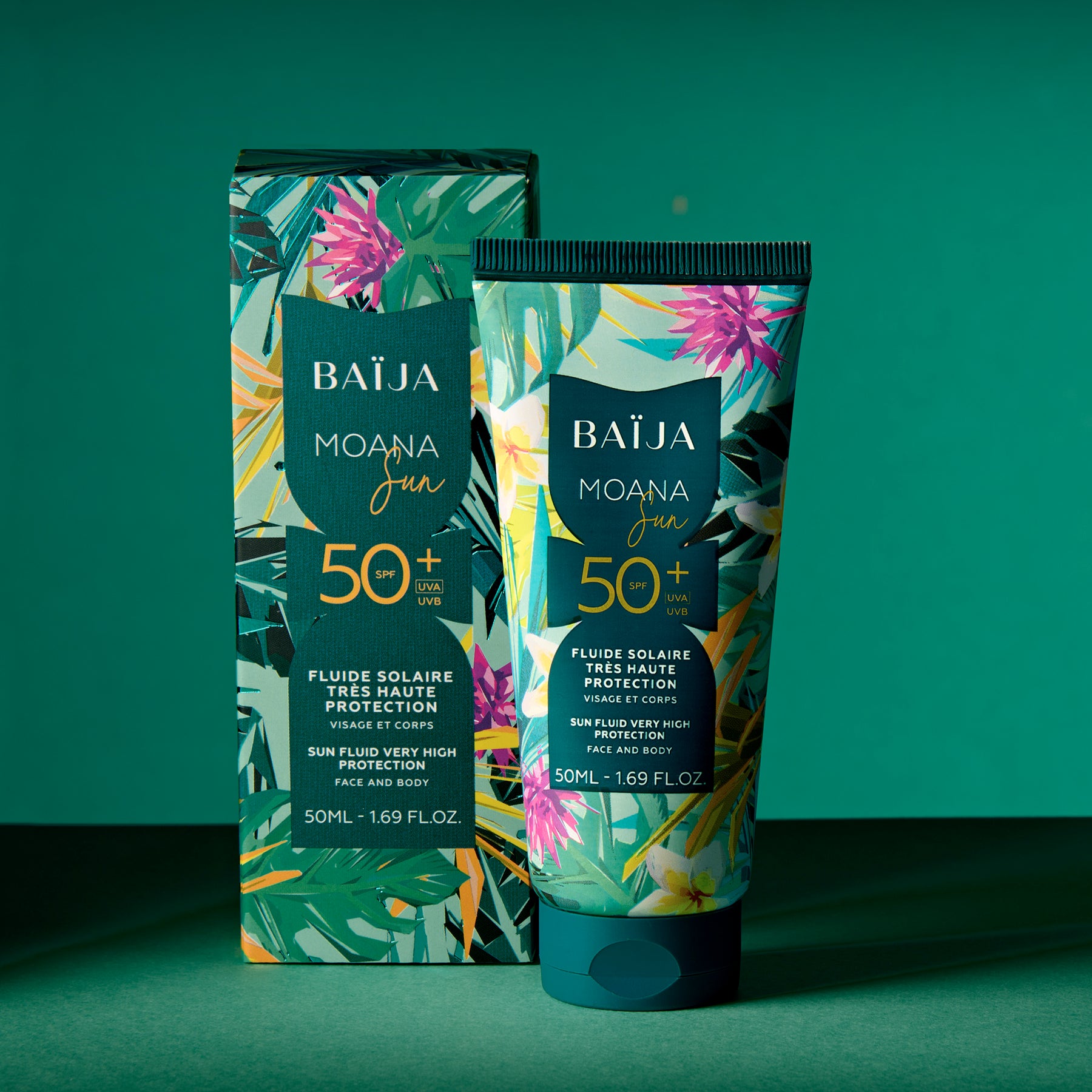 Pack produit sur fond vert/bleu-vert uni : le tube Baïja Moana Sun SPF 50+ est au premier plan, avec son étui/boîte derrière ou à côté. Composition studio, éclairage directionnel avec ombres nettes.