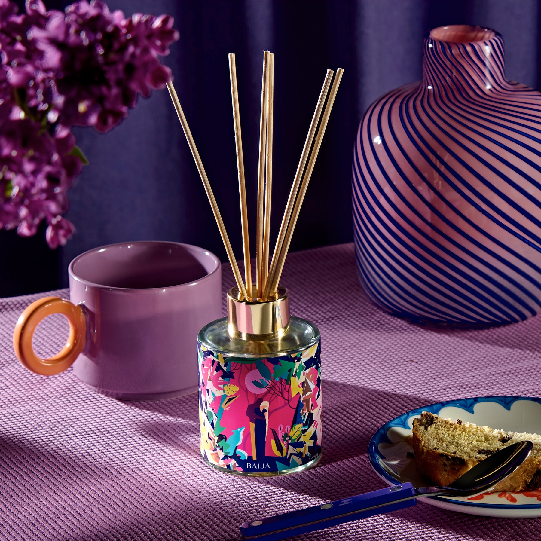 Bouquet parfumé Baïja Minuit Chéri posé sur une table violette, entouré d’une tasse, d’un vase design et d’une part de gâteau, évoquant une atmosphère cocooning et raffinée aux notes boisées et enveloppantes.