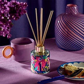 Bouquet parfumé Baïja Minuit Chéri posé sur une table violette, entouré d’une tasse, d’un vase design et d’une part de gâteau, évoquant une atmosphère cocooning et raffinée aux notes boisées et enveloppantes.