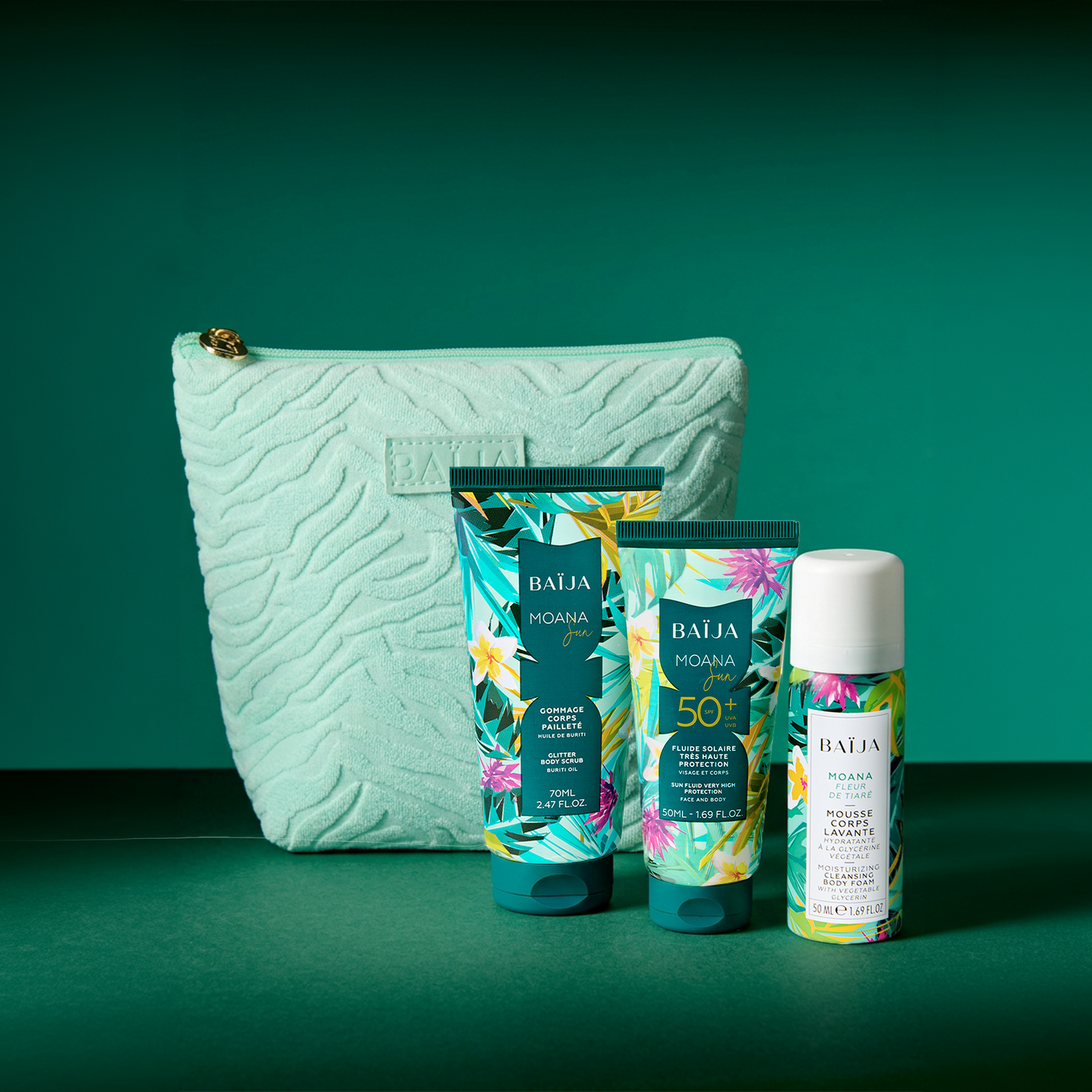 Packshot sur décor bleu profond : trois produits BAÏJA “Moana” (un tube gommage, une mousse corps en flacon, un tube solaire SPF 50+) posés sur un support brillant avec reflets. Des blocs de glace entourent les produits. À droite, une grande trousse vert menthe texturée avec logo “BAÏJA”. Éclairage studio, ambiance fraîche/estivale.