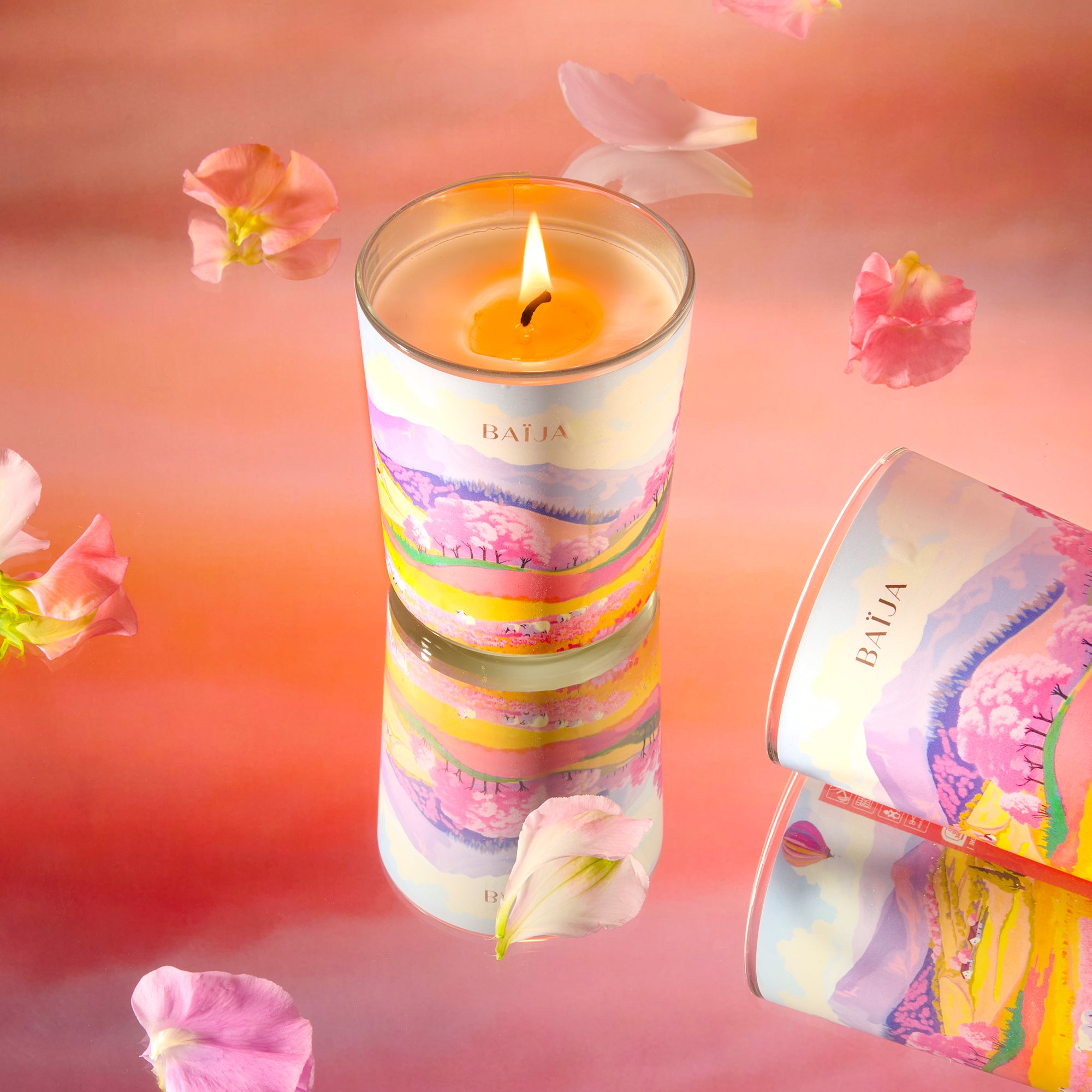 Bougie parfumée Sweet Sunday Baïja allumée, décorée d’un paysage printanier et entourée de pétales délicats sur fond rosé.
