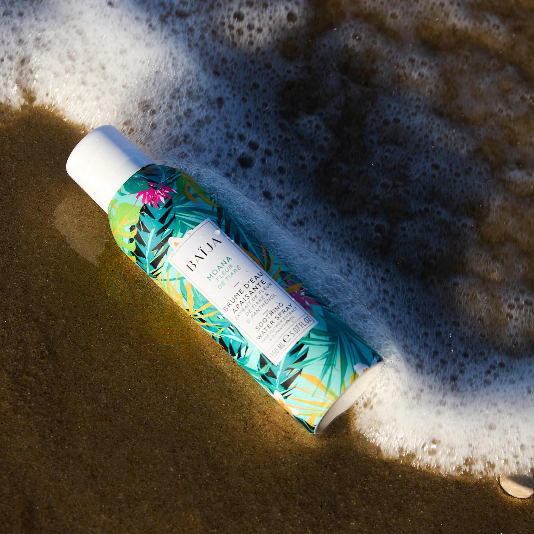 Brume d’eau apaisante Baïja Moana Fleur de Tiaré déposée sur le sable au bord des vagues pour rafraîchir et apaiser la peau.