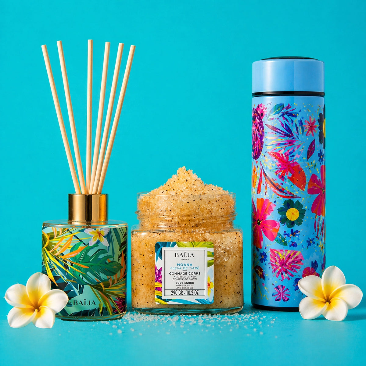 Photo studio sur fond turquoise uni. Au premier plan, trois produits Baïja sont alignés. À gauche, un diffuseur de parfum à bâtonnets : flacon carré en verre avec capot doré, habillé d’un motif tropical vert/jaune, avec des tiges en bois claires qui dépassent vers le haut. Au centre, un pot en verre transparent rempli d’un gommage corps granuleux beige doré ; l’étiquette indique “BAÏJA – MOANA – Fleur de Tiaré” et “Gommage corps”. À droite, une gourde isotherme haute avec un bouchon bleu clair et un habilla